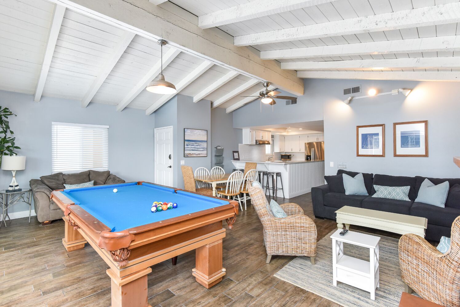 Een ruime woonkamer met een pooltafel, een donkere bank, rieten stoelen en een keuken op de achtergrond.
