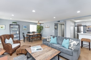 A modern living room with a grey sofa, wicker chairs, a wooden coffee table, and a dining area with a wooden table and chairs.