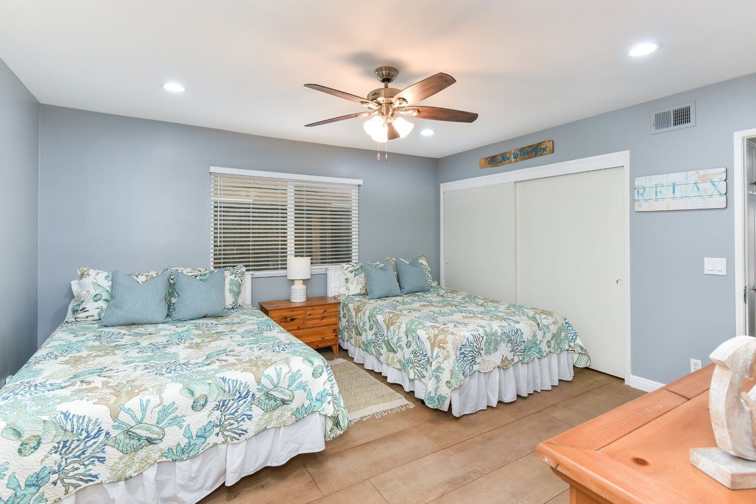 A bedroom with two beds, a ceiling fan, a window with blinds, a wooden nightstand, and a door.