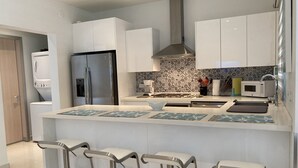 Una cocina moderna con armarios blancos, un refrigerador de acero inoxidable, una estufa a gas y un microondas. Hay tres taburetes de bar con estructura metálica y asientos blancos.