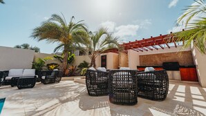 Un área junto a la piscina con mobiliario de mimbre, palmeras y una pérgola.
