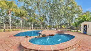 A swimming pool with a brick border, surrounded by trees and a fenced area. - Moama Riverfront Escape: Pool, Fun + Space for Everyone! (Moama)
