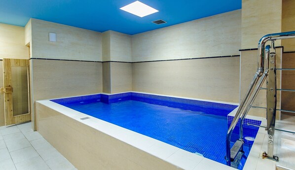Une piscine intérieure moderne, avec un sol carrelé de bleu et un bordure blanche, entourée de murs de couleur beige et d’un plafond bleu.