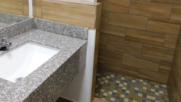 A bathroom with a granite sink, a shower with a checkered floor, and wooden wall tiles. - Hillcrest Motel (Eagle Pass)