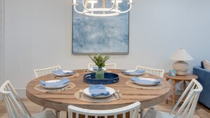 Un comedor con una mesa redonda de madera, sillas blancas, una araña, un florero con follaje y un cuadro abstracto azul en la pared.