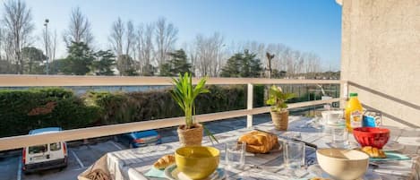 A balcony with a table set for breakfast, including croissants, bowls, and glasses, overlooking a parking area and trees.