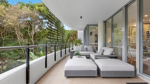 Ein moderner Balkon mit einer grauen Couch und Hocker, einer Glastrennwand und Blick auf Grünflächen.