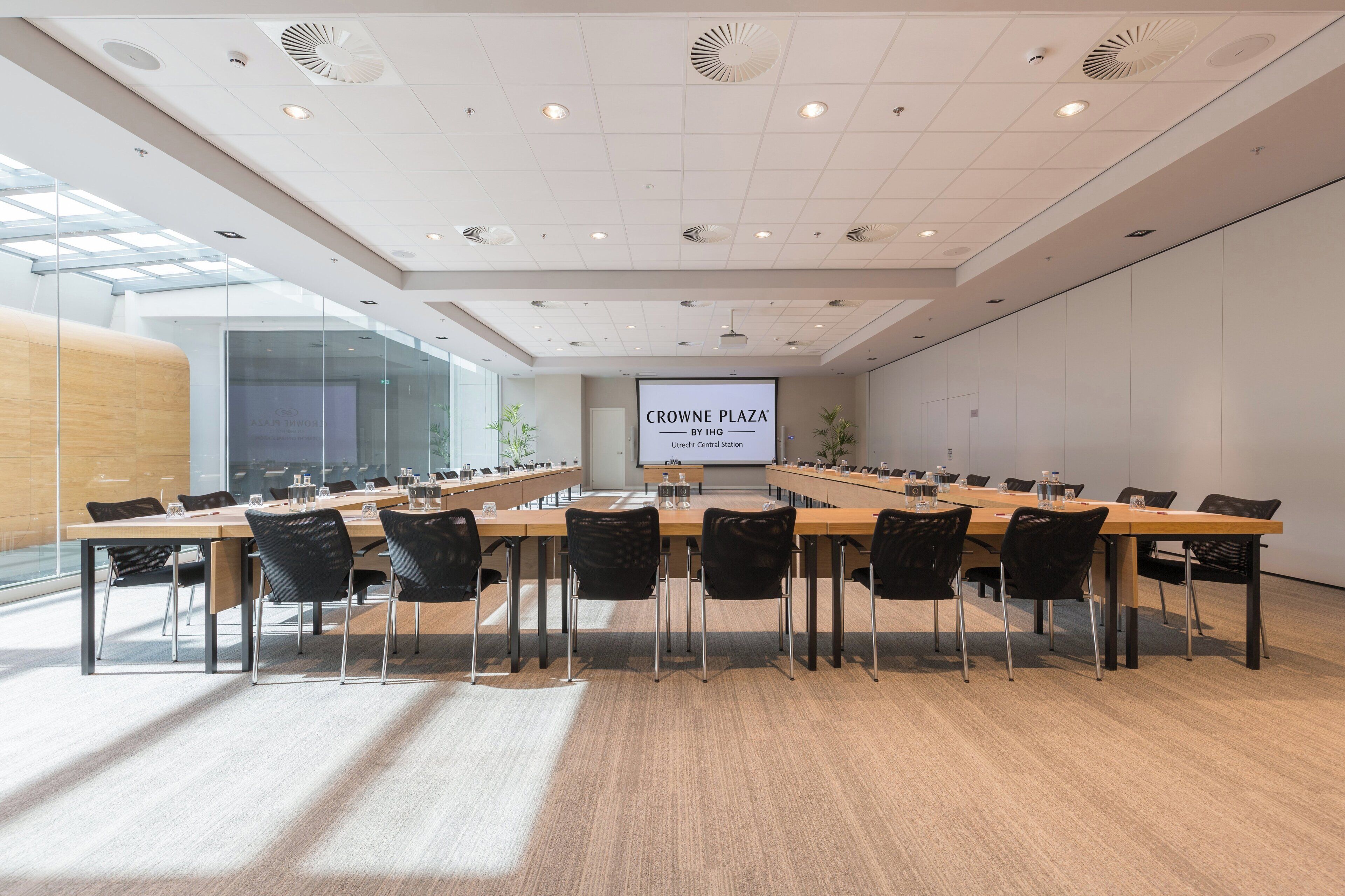 A conference room with a long table, black chairs, a projector screen, and a glass wall on one side.