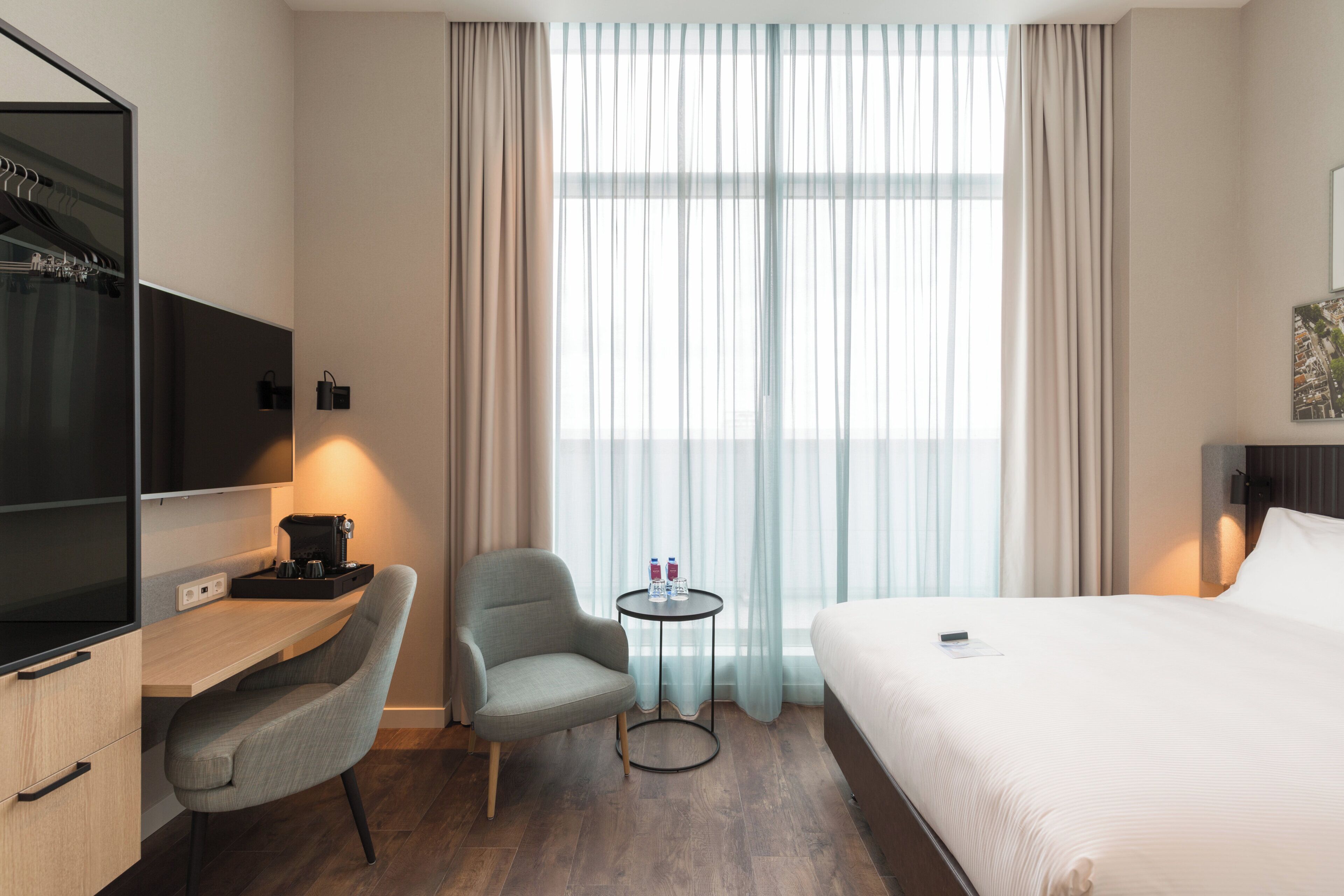 Habitación de hotel con cama, escritorio con cafetera, silla, pequeña mesa con botellas de agua y televisor.