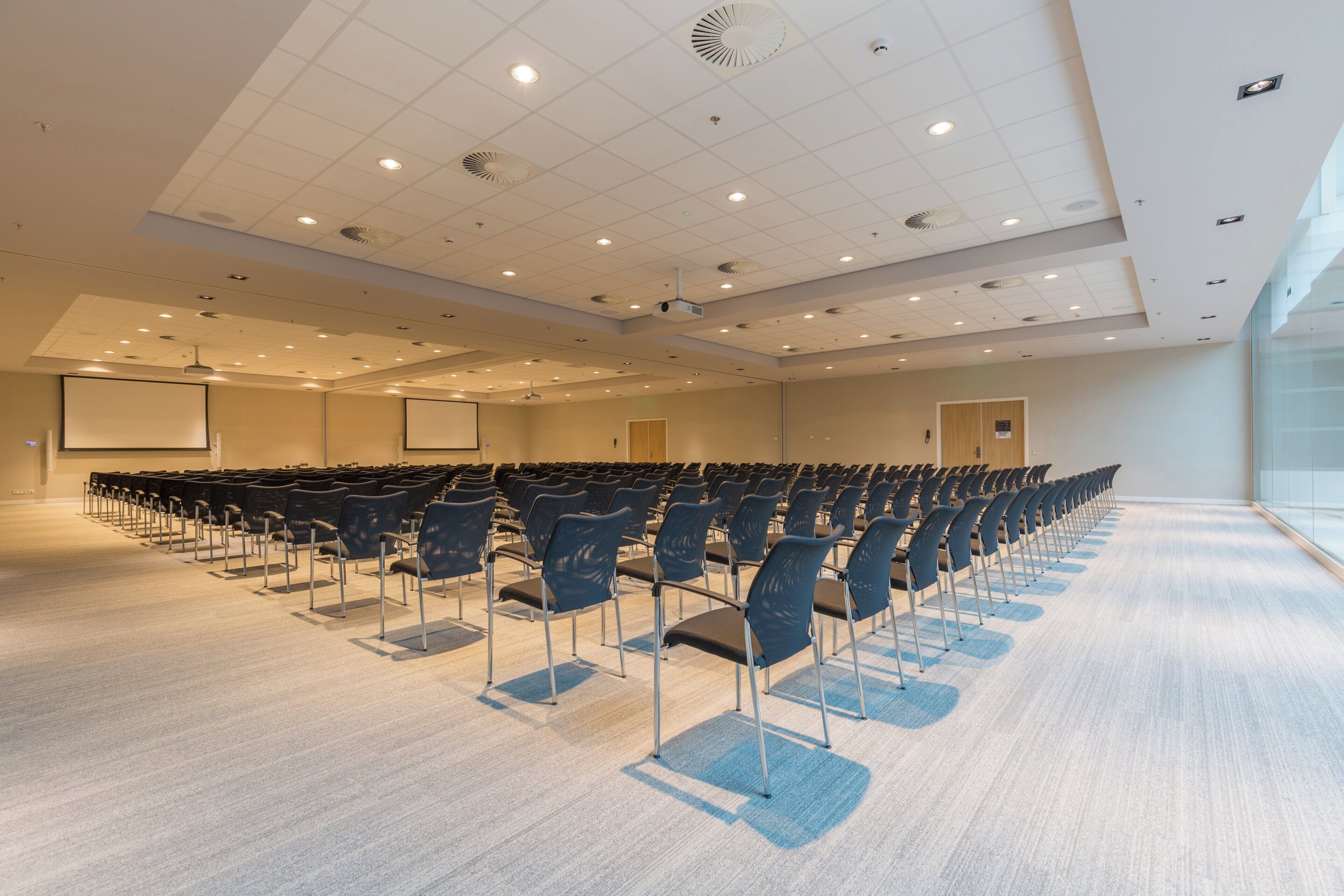 Un salón de conferencias con filas de sillas, una pantalla de proyector y un ventanal grande.