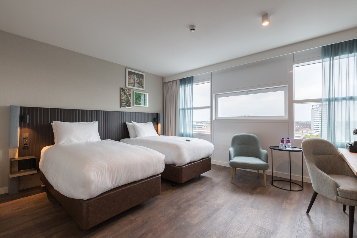 A hotel room with two beds, a desk, a chair, and a view of the city.