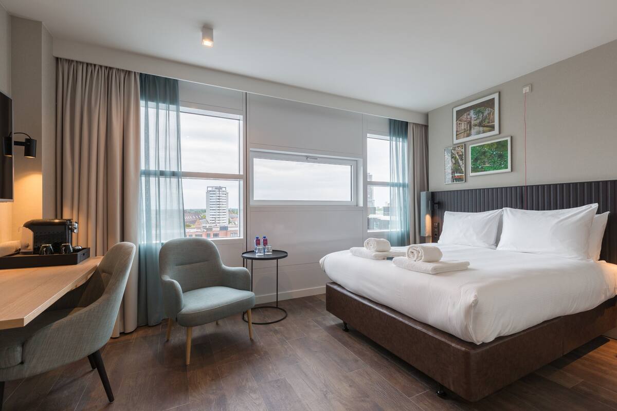 Habitación de hotel con una cama grande, un escritorio con una silla, una pequeña mesa con una cafetera y vistas a la ciudad a través de amplias ventanas.