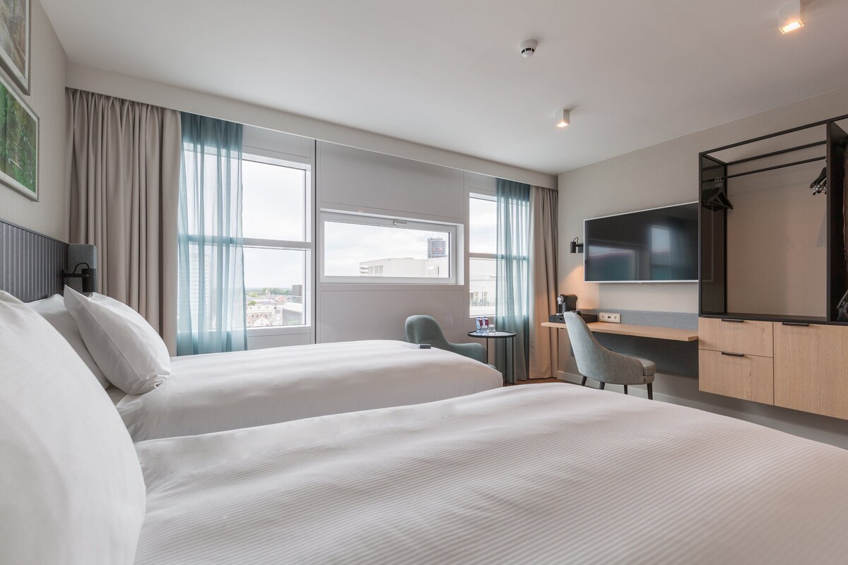 A hotel room with two beds, a desk, a chair, a TV, and a window with curtains.