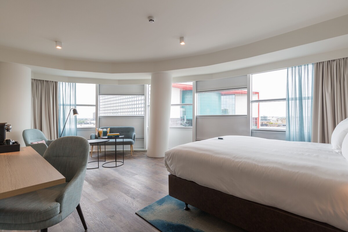 A modern hotel room with a large bed, a desk, and a chair. There are large windows with curtains, a view of the city, and a cityscape visible through the windows.