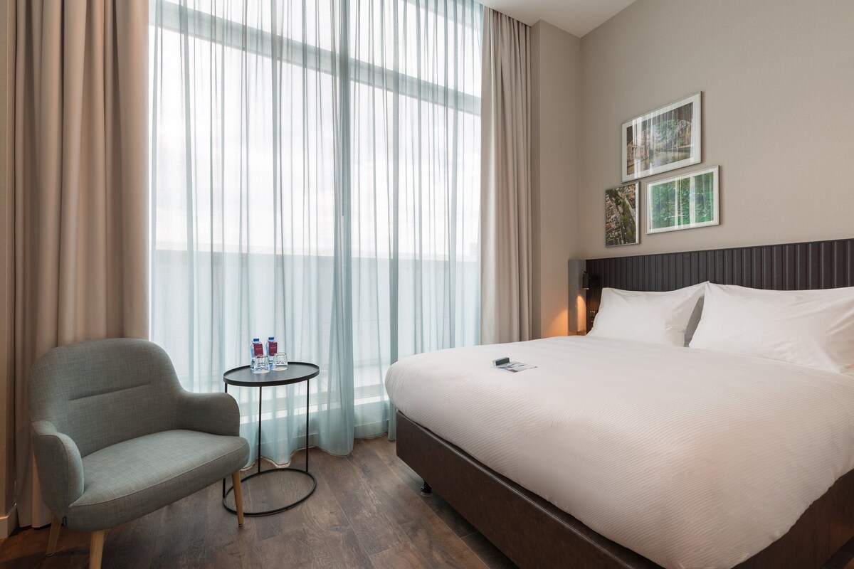 A hotel room with a large bed, a chair, a small table with drinks, and sheer curtains.