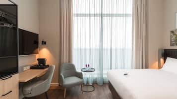 Ein Hotelzimmer mit Bett, Schreibtisch mit Stuhl, einem kleinen Tisch mit zwei Flaschen, einem Fernseher und einem Fenster mit Vorhängen.
