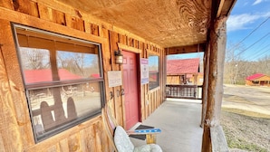 A wooden building with a porch, a red door, and a window with a view of a red-roofed building in the background. - Sylamore Depot Cabin # 302 (Mountain View)
