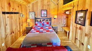 A wooden cabin bedroom with a bed, two bedside tables, a lamp, a framed picture, and a shelf with a suitcase. - Sylamore Depot Cabin # 302 (Mountain View)