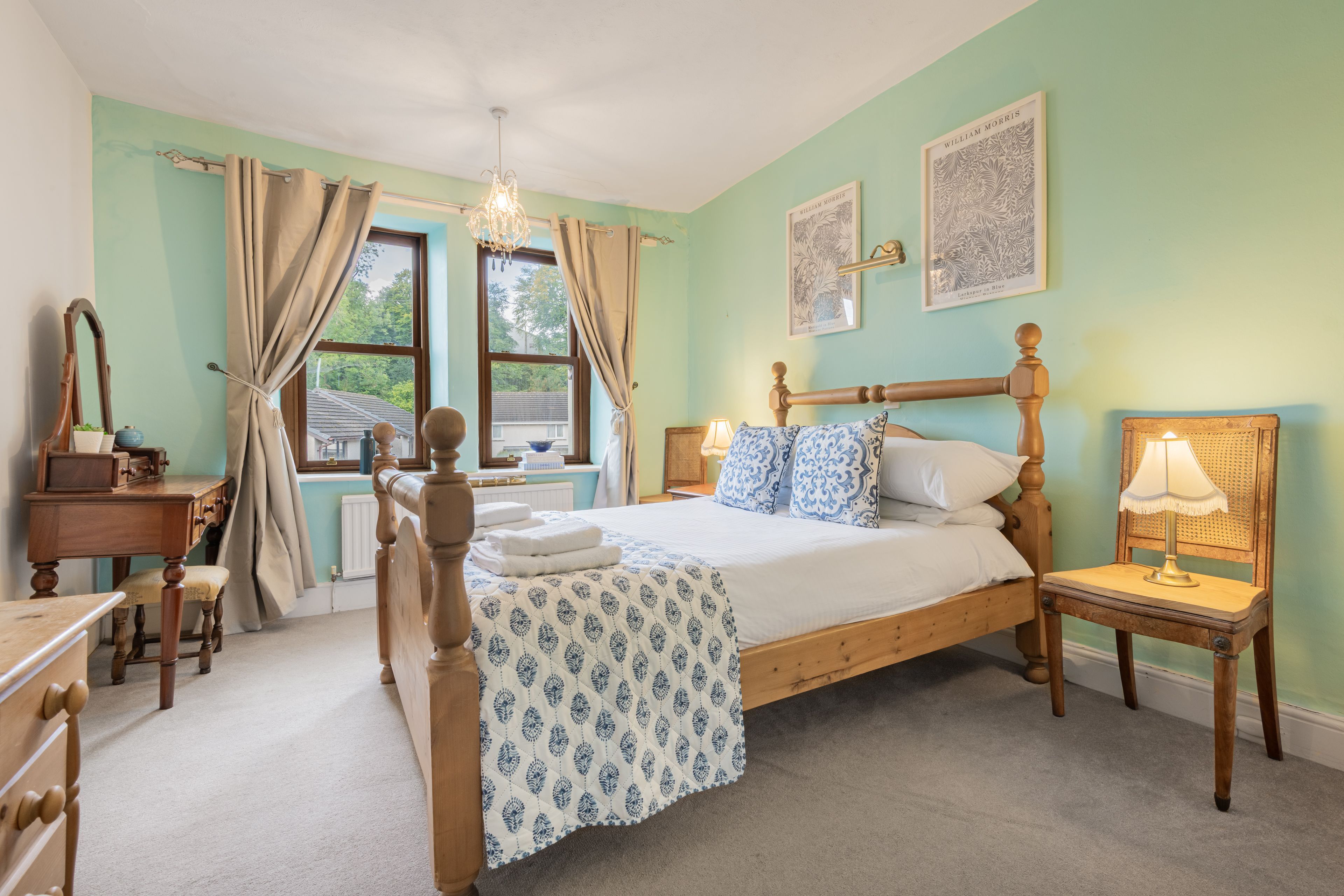 A bedroom with a wooden bed, a desk, a chair, a lamp, and two framed pictures on the wall.