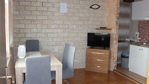 Un espacio habitable compacto con pared de ladrillo, una mesa de comedor de madera con dos sillas, un televisor sobre un mueble de madera y una zona de cocina con armarios y un fregadero.