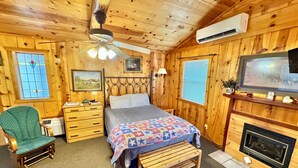 A cozy wooden cabin bedroom with a bed, a chair, a dresser, a fireplace, and a ceiling fan. - Liberty Cabin Number 301 (Mountain View)
