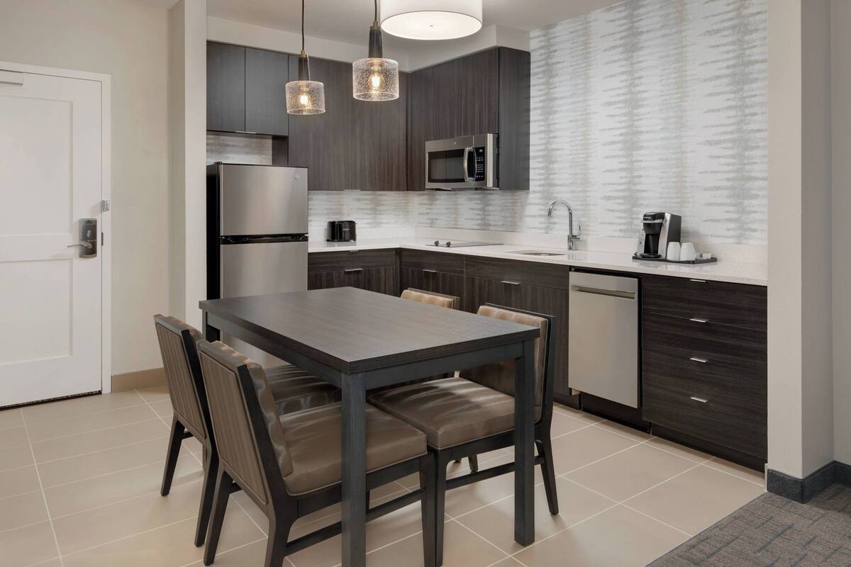 A modern kitchen with dark cabinetry, a stainless steel refrigerator, a microwave, and a coffee maker. There is a dining table with chairs and a light fixture above it.