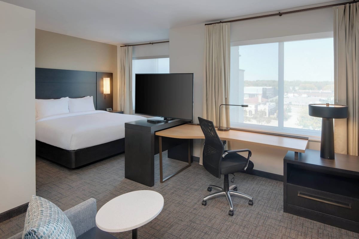 A hotel room with a bed, desk, chair, TV, and a view of the city.