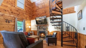 A cabin interior with a fireplace, wooden walls, a sofa, a chair, and a spiral staircase. - ER24 - Honeysuckle Holler | Great Location..Close to town (Pigeon Forge)