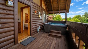 A wooden cabin with a hot tub on the deck, a door leading to a room, and a view of greenery. - ER24 - Honeysuckle Holler | Great Location..Close to town (Pigeon Forge)