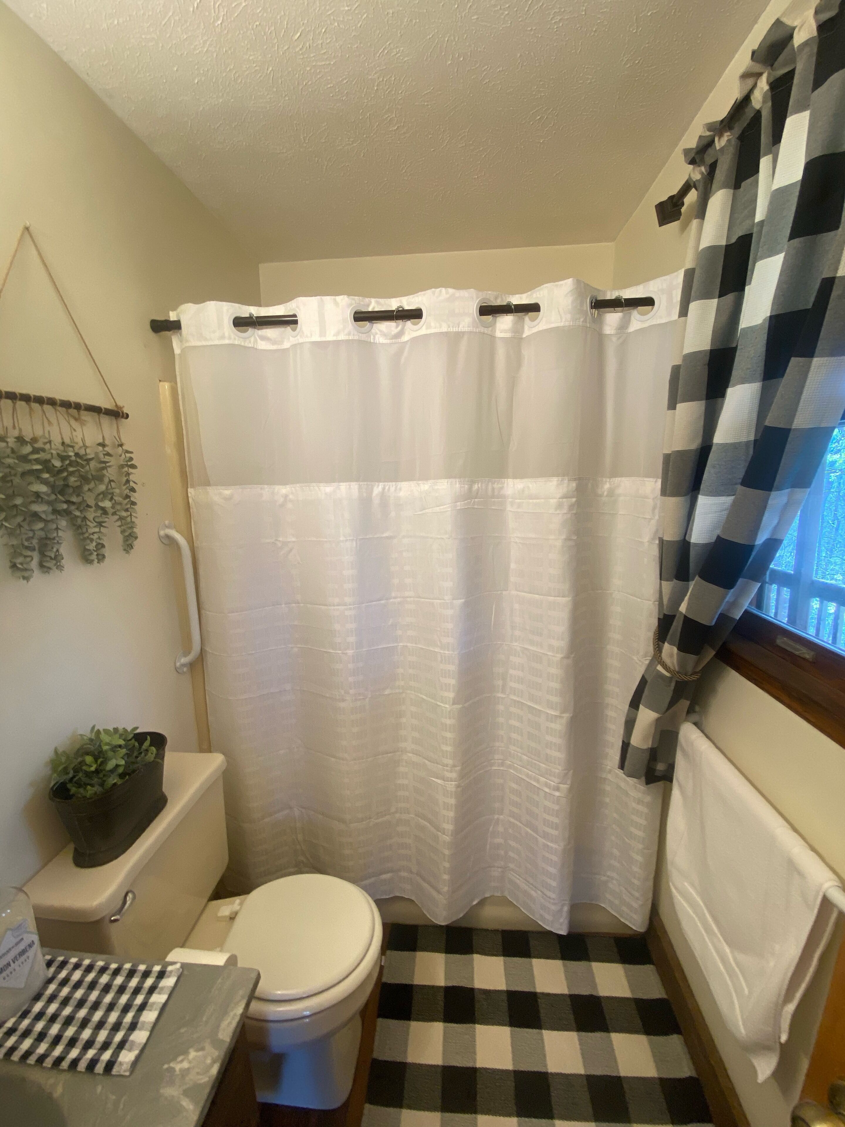 Une petite salle de bain avec des toilettes blanches, un carrelage noir et blanc, un rideau de douche blanc et une fenêtre ornée d’un rideau à carreaux.