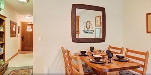 A dining area with a wooden table set for four, a mirror on the wall, and a shelf with various items.
