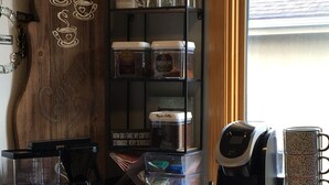 Eine Küchenarbeitsplatte mit Kaffeemaschine, Kaffeemühle und Zubehör. Eine Ablagefläche mit Gläsern und einem Schild mit der Aufschrift: 'Wie nehme ich meinen Kaffee?'
