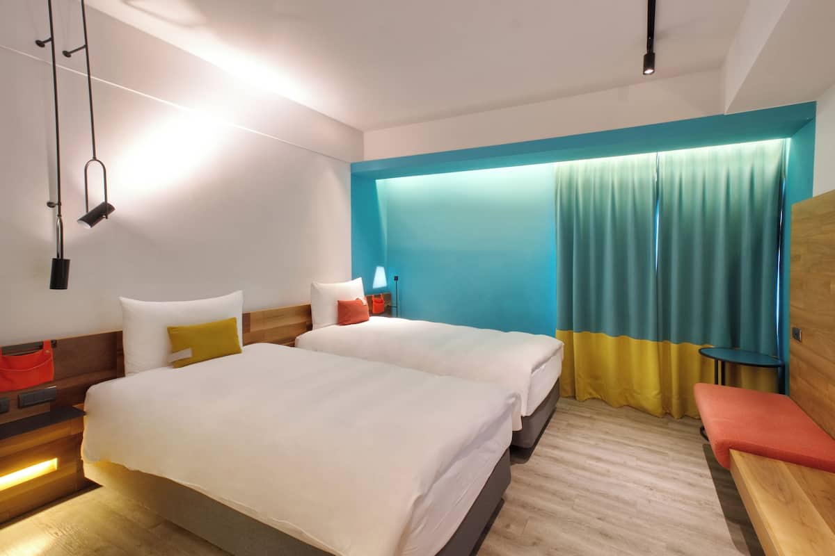 A modern hotel room with two beds, a wooden bench, a small table, and a large window with curtains.