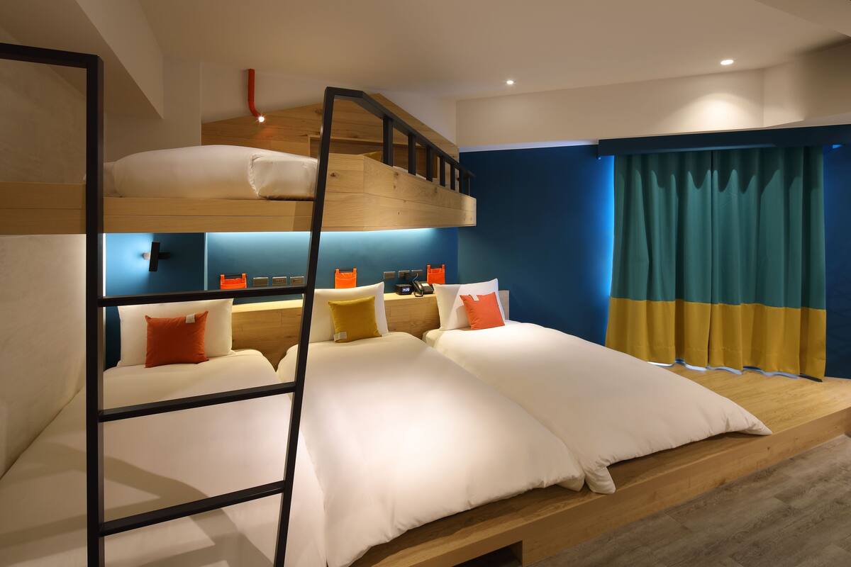 A bunk bed room with a wooden ladder, two beds with white and orange bedding, a desk with a chair, and a window with curtains.