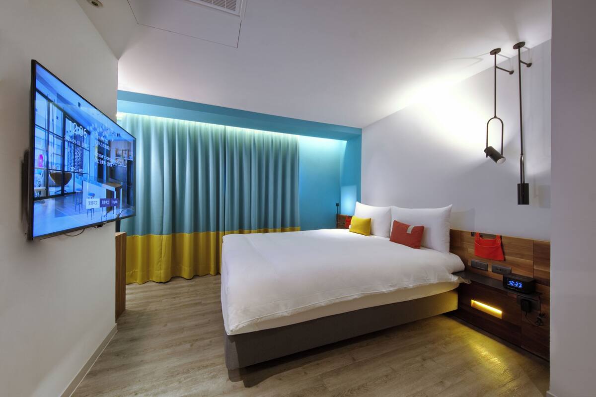 A modern hotel room with a large bed, a flat-screen TV mounted on the wall, a bedside table with a digital clock, and a window with curtains.