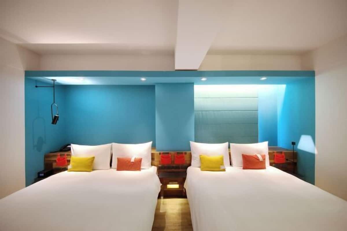 A hotel room with two beds, each with white linens and colorful pillows. There is a wooden headboard, a nightstand with a lamp, and a window with a view.