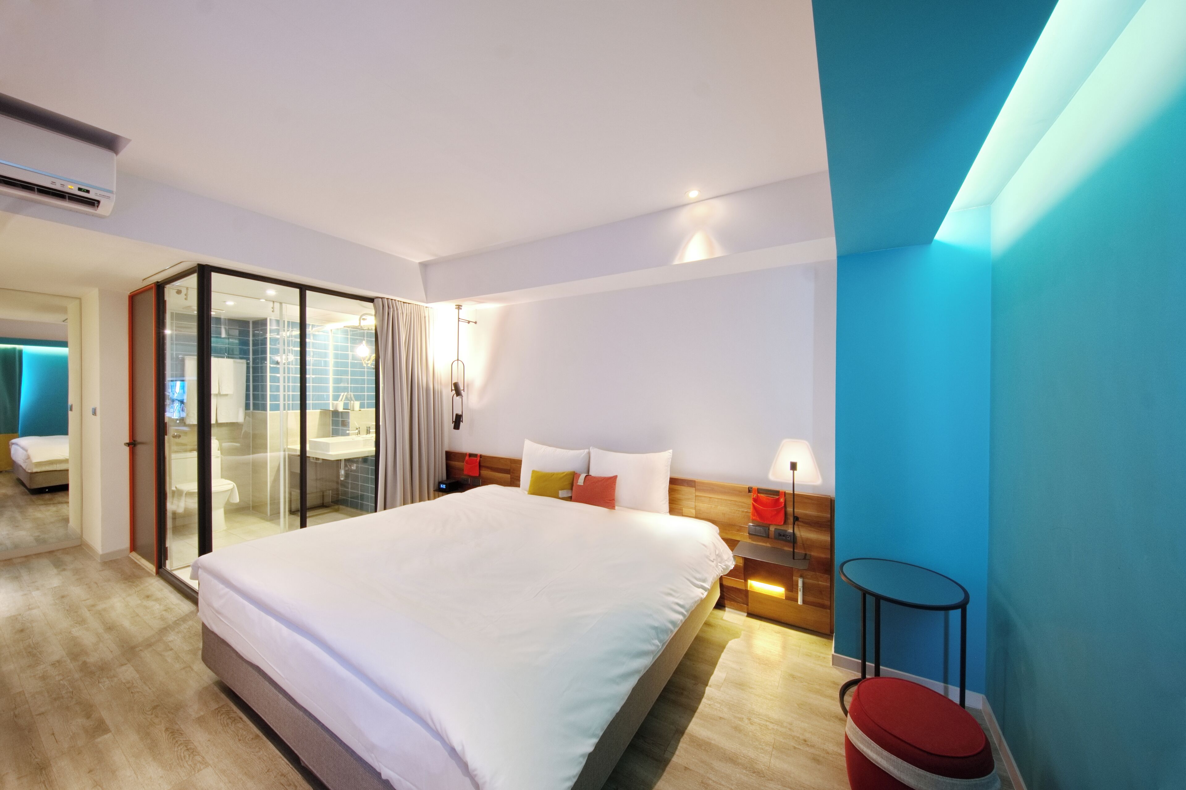 Una habitación de hotel moderna con una cama grande, un ventanal que da a un balcón, una mesita de noche de madera y una pequeña mesa.
