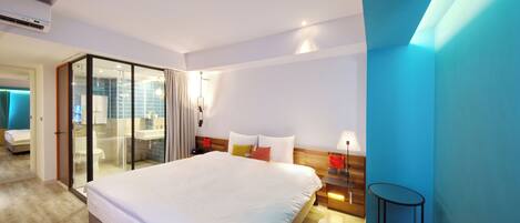 Una habitación de hotel moderna con una cama grande, un ventanal que da a un balcón, una mesita de noche de madera y una pequeña mesa.