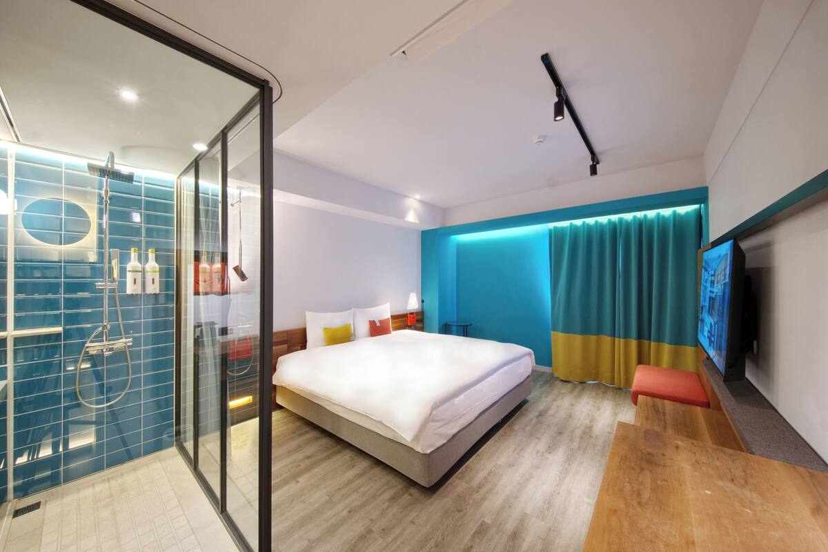 A modern hotel room with a large bed, a glass shower enclosure, a flat-screen TV, and a wooden bench.