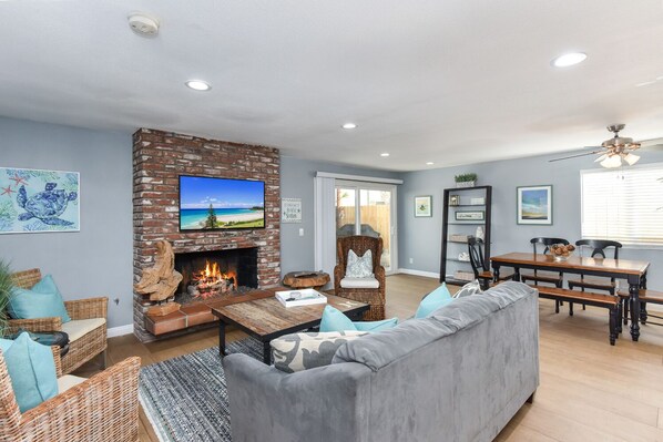 A living room with a fireplace, a television, wicker furniture, a gray sofa, and a dining area with a table and chairs.