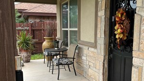 Un porche cubierto con un muro de piedra, una puerta negra adornada con un arreglo floral y una mesa de patio con sillas.