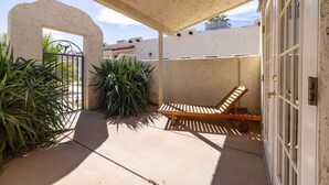 A covered patio area with a wooden lounge chair, a metal gate, and potted plants.