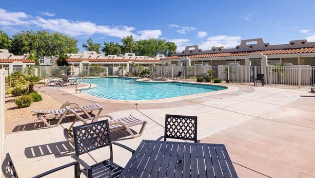 A swimming pool with lounge chairs and a table, surrounded by a fence and buildings.