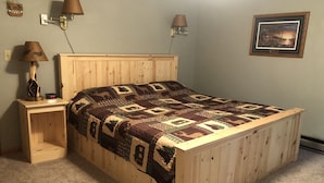 A wooden bed with a patterned blanket, two bedside lamps, a small table with a clock, and a framed picture on the wall.