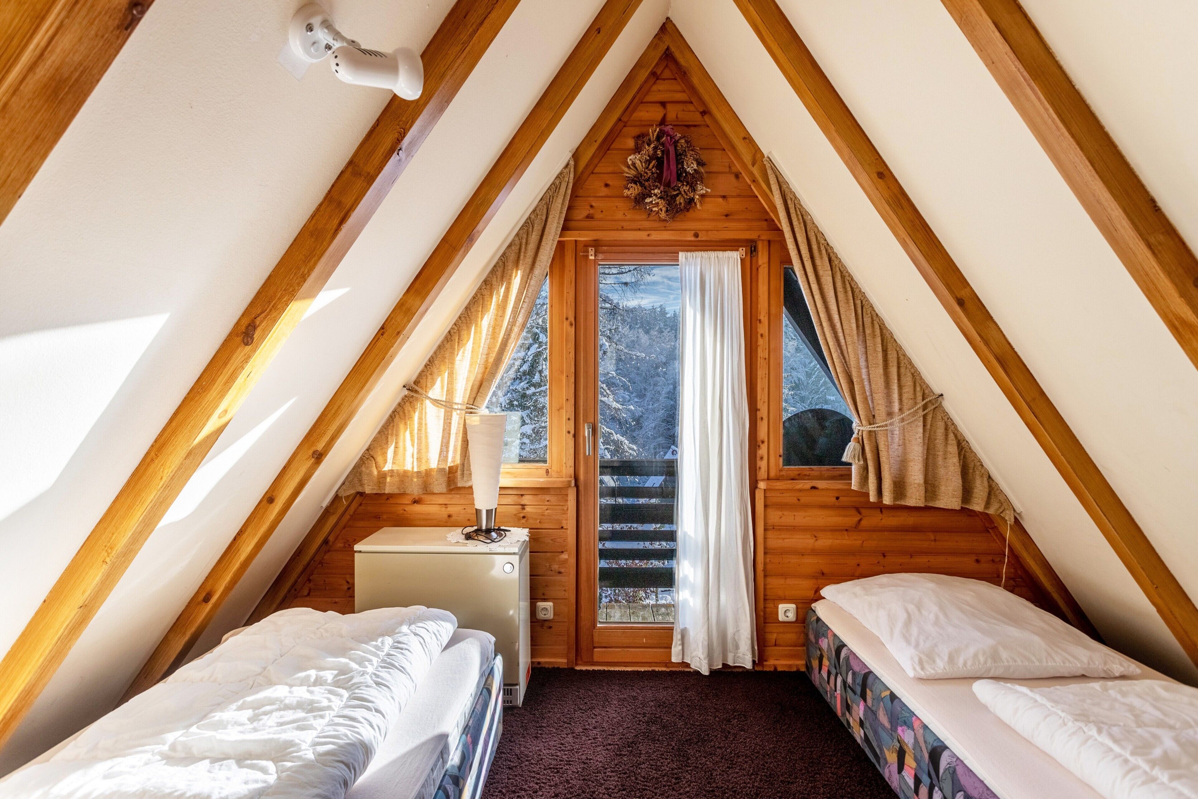 A small, cozy attic bedroom with a sloped ceiling, two beds, a small desk, and a window with curtains.
