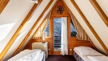 A small, cozy attic bedroom with a sloped ceiling, two beds, a small desk, and a window with curtains.