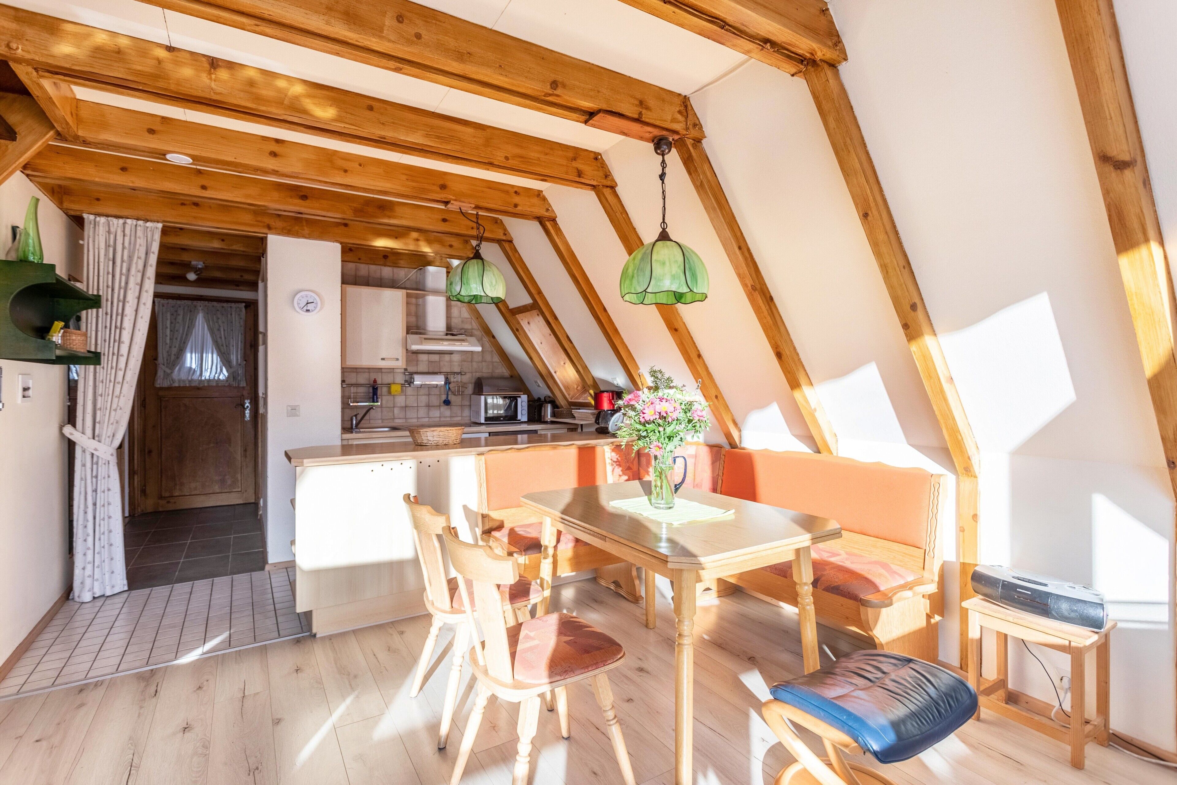 A cozy attic bedroom with a wooden ceiling, a dining table with chairs, a bed, and a small table with a lamp.