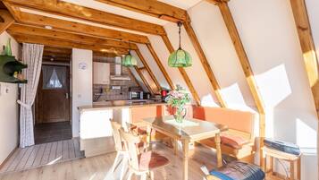 A cozy attic bedroom with a wooden ceiling, a dining table with chairs, a bed, and a small table with a lamp.