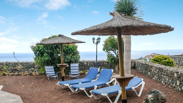 Une terrasse en bord de mer, dotée de chaises longues, de parasols en chaume et d’un mur de pierre d’où l’on peut admirer la mer.