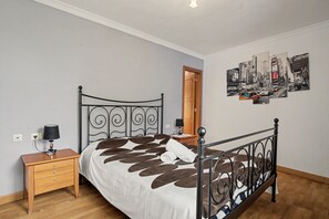 A bedroom with a bed, a nightstand, a lamp, a wooden dresser, and a wall with a cityscape artwork.